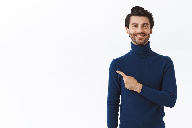 homme avec des poils dans un pull à col haut élégant, pointant fièrement vers la gauche et regarde la caméra, recommande la promo, annonce le produit