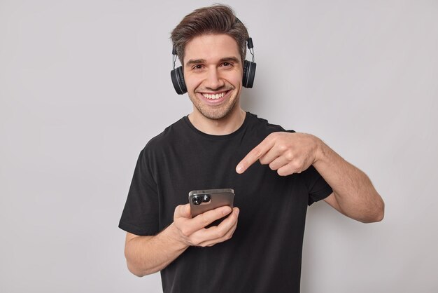 l'homme pointe vers l'écran du smartphone porte des écouteurs sans fil sur les oreilles s'habille en t-shirt noir décontracté isolé sur blanc montre à quelle vitesse ses nouveaux gadgets fonctionnent.