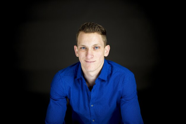 Homme portant un t-shirt bleu et souriant à la caméra avec un mur noir