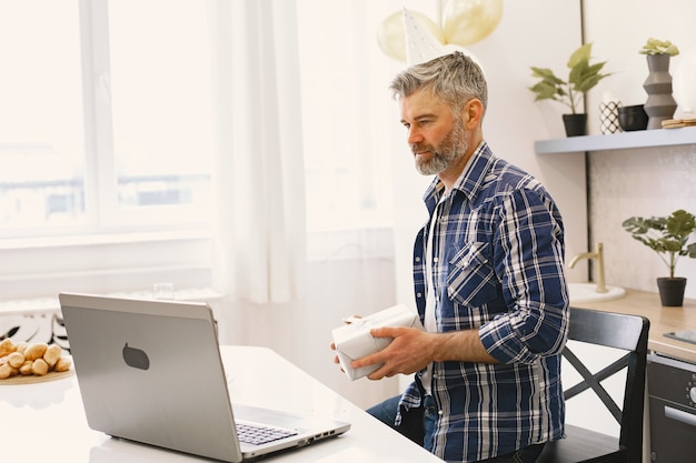 L'homme porte un chapeau de fête. L'homme garde une boîte avec un cadeau. L'homme a un appel via son ordinateur portable.