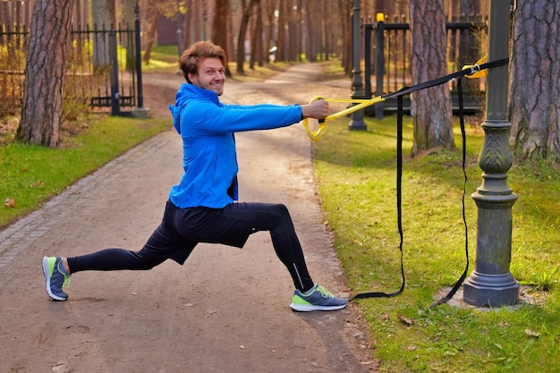 Homme positif attrayant dans un imperméable bleu faisant de l'exercice dans un parc avec des bandes de fitness trx.