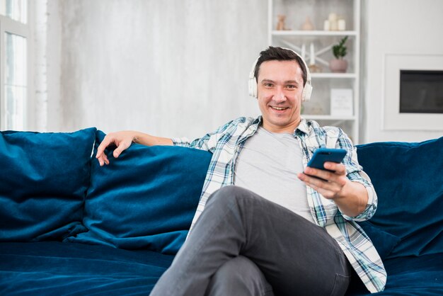 Homme positif écoute de la musique au casque et tenant un smartphone sur un canapé dans la chambre