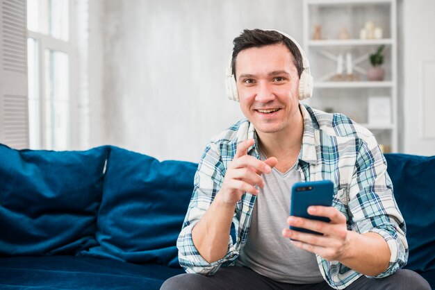 Homme positif écoute de la musique au casque et tenant un smartphone sur un canapé