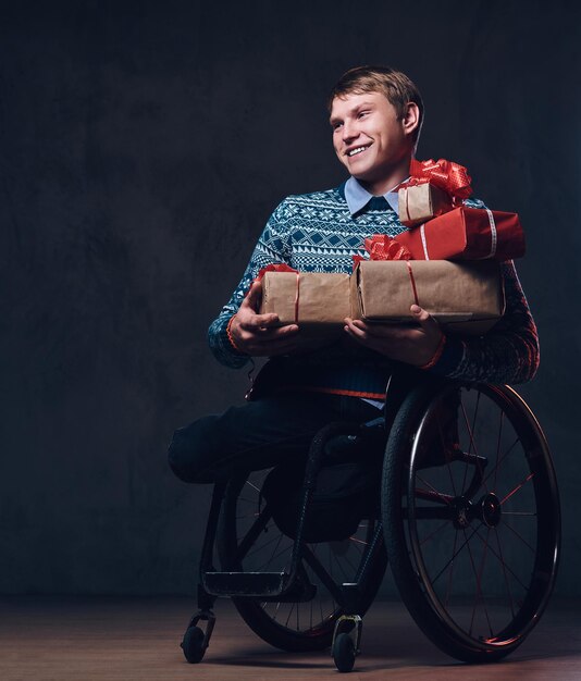 Homme positif en fauteuil roulant avec des cadeaux de Noël sur fond gris foncé.