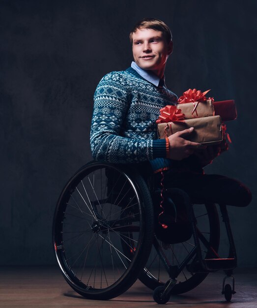 Homme positif en fauteuil roulant avec des cadeaux de Noël sur fond gris foncé.