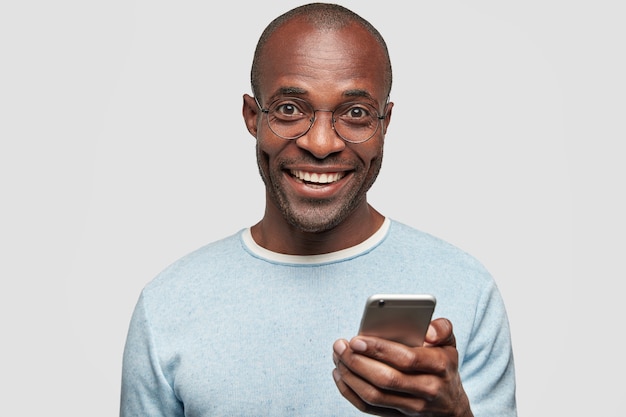 Homme positif avec un large sourire, détient un téléphone portable moderne, tape des messages texte et des commentaires, surfe sur les réseaux sociaux