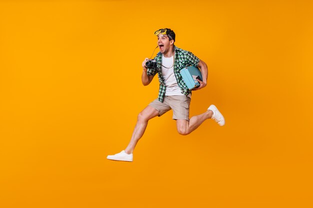 Un homme positif avec un masque de plongée sur la tête prend des photos avec un appareil photo rétro. Guy en short et chemise verte sautant avec valise sur l'espace orange.