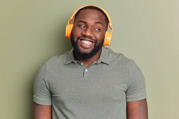 L'homme positif avec des sourires à pleines dents écoute la piste audio préférée via des écouteurs détourne les yeux pensivement habillés en t-shirt décontracté pose contre un mur vert foncé