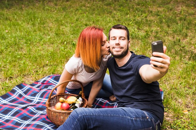 Homme prenant selfie par téléphone portable avec sa petite amie l&#39;embrassant sur la joue au pique-nique