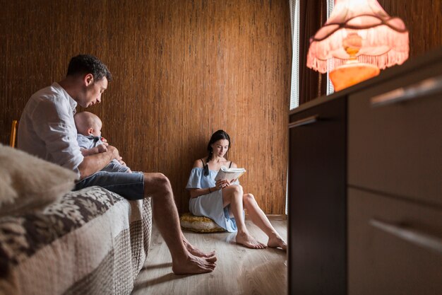 Homme prenant soin de son bébé près de la femme lisant un livre à la maison