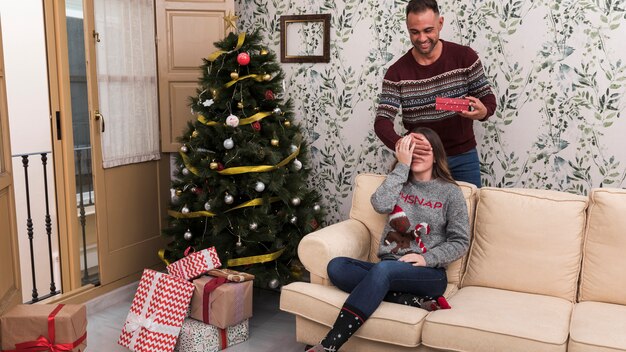 Homme présentant une boîte-cadeau et fermant les yeux sur une femme assise sur un canapé près de l&#39;arbre de Noël