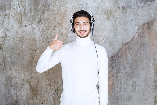 Homme en pull blanc portant des écouteurs et écoutant la musique et montrant un signe de main positif.