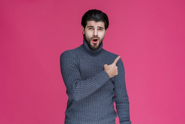 Homme en pull gris pour pointer quelque chose ou présenter quelqu'un à l'aide du doigt pointé.