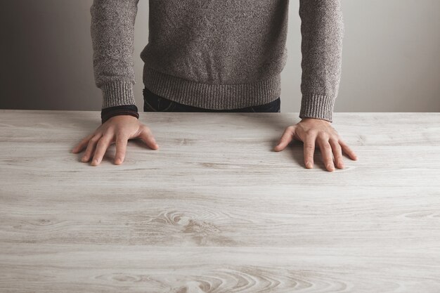 L'homme en pull gris tient la main sur une table en bois blanc vierge, isolée sur un mur simple