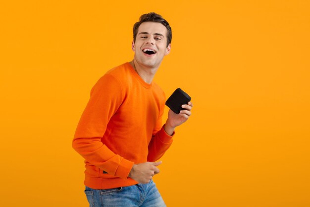 homme en pull orange tenant un haut-parleur sans fil heureux d'écouter de la musique s'amusant isolé sur jaune