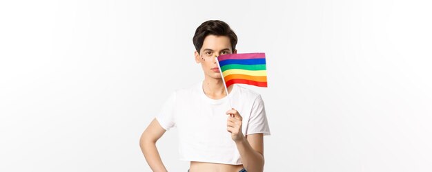 Homme queer attrayant de la communauté lgbtq avec flitter sur le visage agitant le drapeau arc-en-ciel de fierté et regardant c