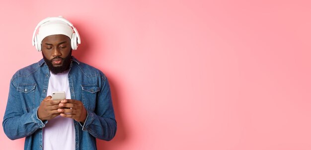 Photo gratuite un homme qui a l'air sérieux en lisant des messages au téléphone en écoutant de la musique dans des écouteurs debout sur du rose