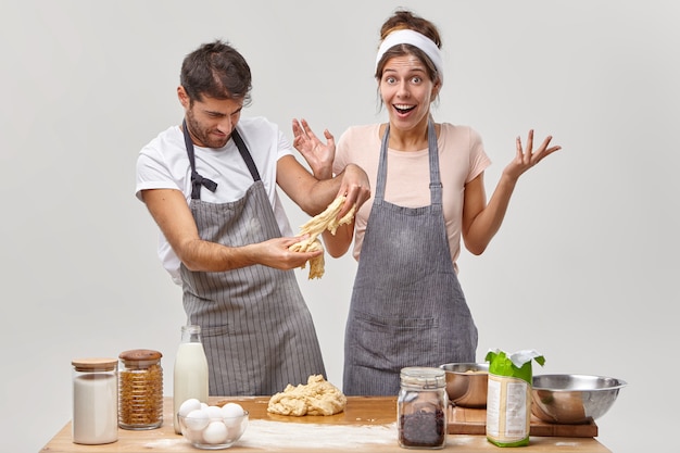 Un homme qui travaille dur en tablier pratique des compétences en pâtisserie avec sa femme, essaie de faire de la pâte pour une pâtisserie ou une tarte, cuit à la maison, pose à la cuisine près de la table avec des ingrédients. Il est temps de préparer un délicieux dîner