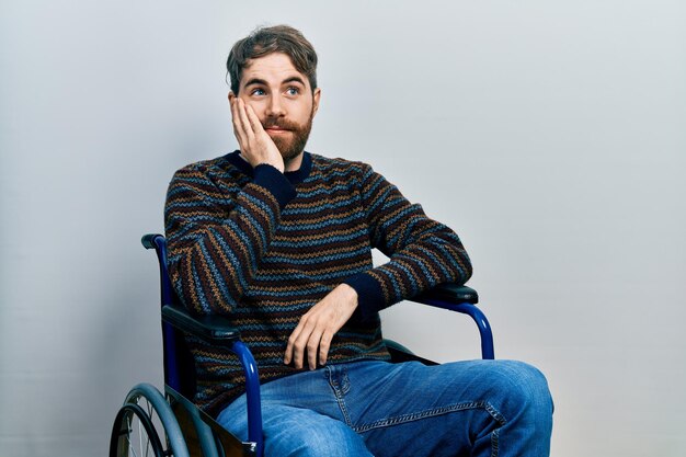 Homme de race blanche avec barbe assis sur un fauteuil roulant à l'air stressé et nerveux avec les mains sur la bouche se ronger les ongles. problème d'anxiété.