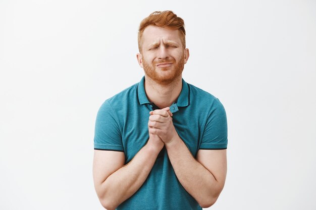 Homme de race blanche rousse avec poils en t-shirt vert, fermant les yeux en fronçant les sourcils, serrant les mains ensemble sur la poitrine tout en mendiant passionnément quelque chose