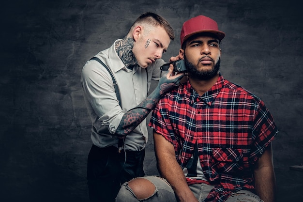 Un homme de race blanche tatoué vêtu d'une chemise blanche coupant la barbe d'hommes hipster noirs vêtus d'une chemise polaire rouge.