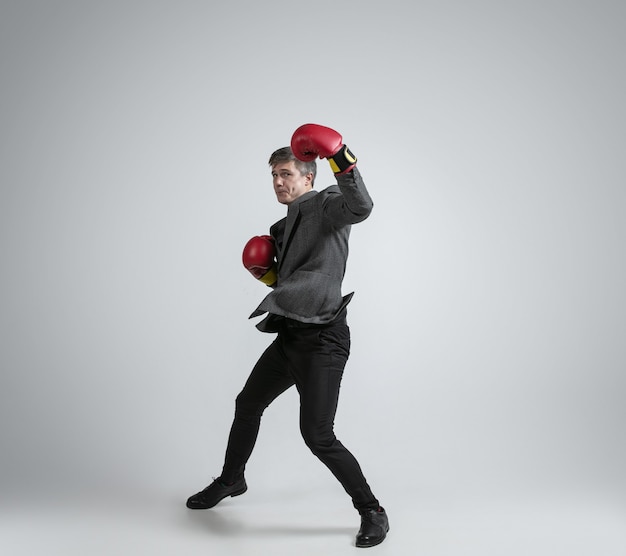 Homme de race blanche en vêtements de bureau boxe avec deux gants rouges sur fond gris.