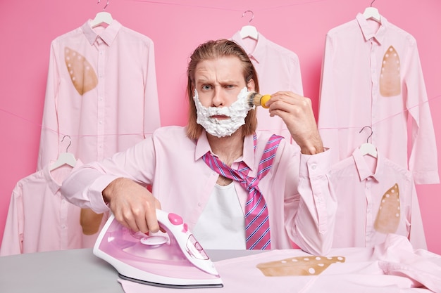 l'homme rase la barbe avec une expression mécontente porte une chemise et une cravate autour du cou utilise un fer électrique à vapeur pour caresser les vêtements se tient près d'une planche à repasser