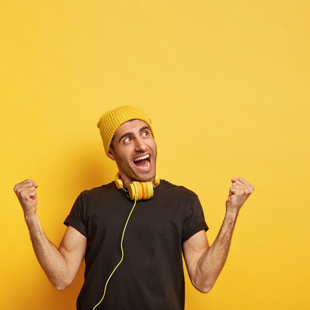 Un homme ravi lève les poings fermés, se sent énergique et optimiste, porte un chapeau jaune et un t-shirt noir, fait des gestes joyeux, écoute de la musique dans des écouteurs