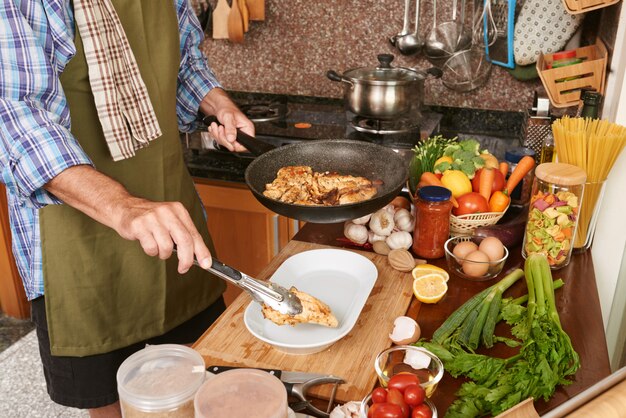 Homme recadré en tablier mettant le poulet frit de la poêle à frire dans l'assiette