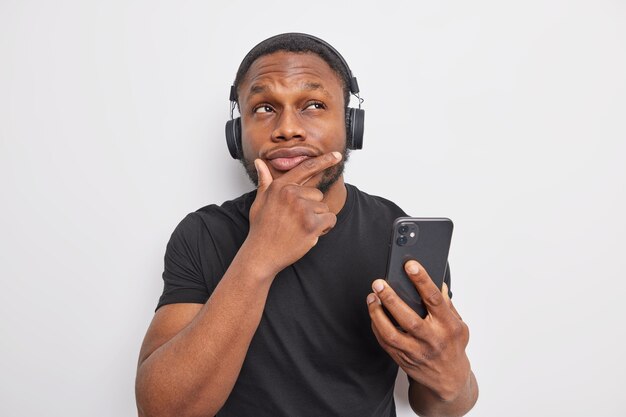 Un homme réfléchi à la peau foncée tient le menton avec une expression pensive utilise un téléphone portable et des écouteurs stéréo pour écouter de la musique se tient pensif à l'intérieur sur fond blanc. Laisse moi y réfléchir