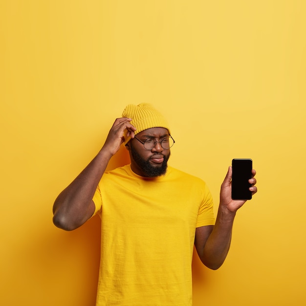 Un homme réfléchi perplexe se gratte la tête, réfléchit à la création d'une nouvelle application, vous montre l'écran du smartphone, vêtu de vêtements jaunes