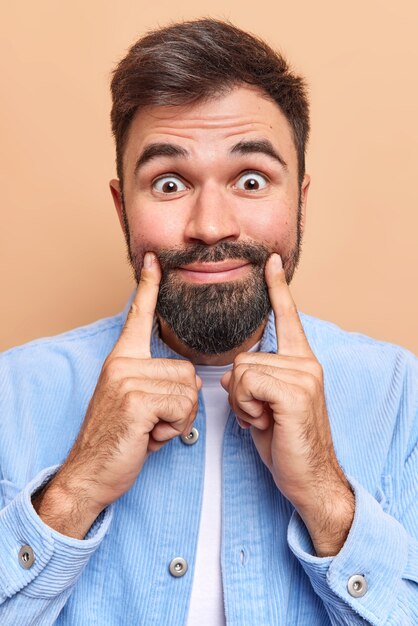 l'homme regarde étonnamment la caméra garde l'index près des coins des lèvres force le sourire vêtu d'une chemise de velours bleu isolée sur marron