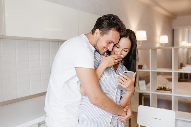 Homme romantique avec une coiffure courte embrassant une dame brune en riant porte un pyjama mignon