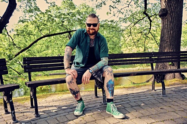 Homme rousse barbu tatoué dans une chemise polaire se reposant sur un banc dans un parc d'été.