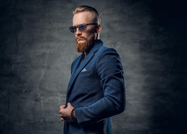 Homme rousse barbu vêtu d'un costume bleu et de lunettes de soleil sur fond gris foncé.