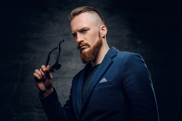 Homme rousse barbu vêtu d'un costume bleu et de lunettes de soleil sur fond gris foncé.