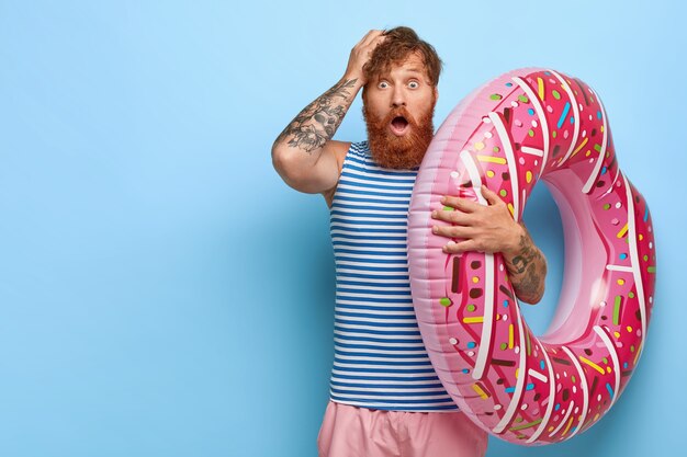 homme rousse effrayé recrée pendant les vacances d'été, tient un anneau de bain rose, choqué d'oublier ce qui est nécessaire pour se reposer, porte un gilet de marin rayé et un short garde la main sur la tête. Concept Omg