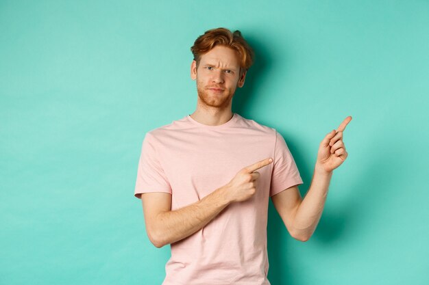 Homme rousse sceptique et douteux en t-shirt pointant les doigts dans le coin supérieur droit, montrant une offre promotionnelle avec un visage mécontent, debout sur fond turquoise.