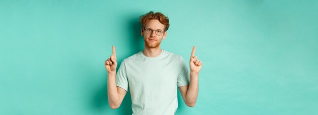 Homme rousse sceptique et non amusé dans des verres et un t-shirt pointant les doigts vers le haut et montrant quelque chose u