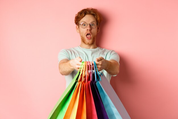 Homme rousse surpris étirer les mains avec des sacs à provisions, vous donner des cadeaux, debout sur fond rose.