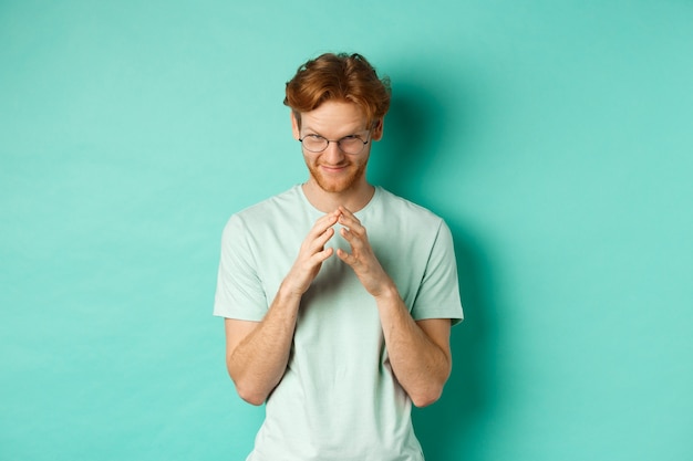 Homme roux sournois dans des lunettes et un t-shirt lançant une idée, des doigts de clocher et un regard sous le front avec un sourire narquois et suffisant, debout sur un fond de menthe.