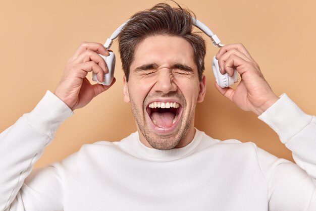 l'homme s'exclame fort garde la bouche grande ouverte enlève des écouteurs stéréo écoute de la musique à volume élevé vêtu d'un pull blanc décontracté isolé sur beige.