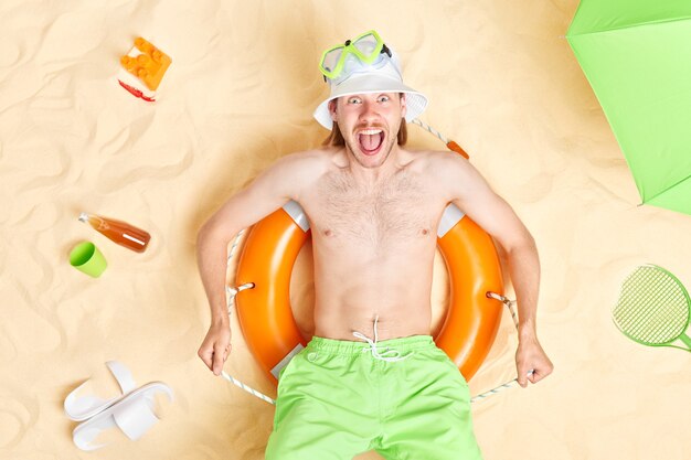 L'homme s'exclame fort garde la bouche largement ouverte pose à la plage porte un short vert panama blanc masque de plongée se trouve sur bouée gonflée