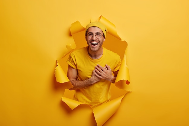 Photo gratuite un homme satisfait se tient dans un trou de papier, garde les mains près du cœur, exprime sa gratitude