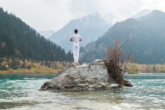 Un homme se tient sur une pierre au centre d'un lac de montagne et pratique le yoga. pose vrikshasana.