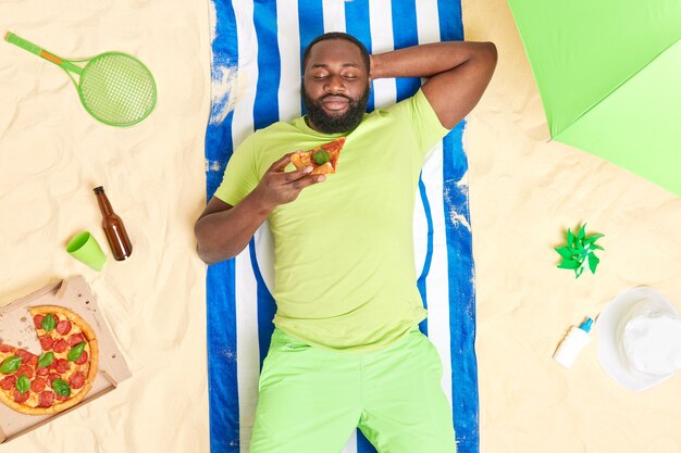 l'homme se trouve à la plage mange de la pizza se repose bien pendant les vacances d'été vêtu d'un t-shirt vert et d'un short pose à une serviette sur le sable avec divers articles autour