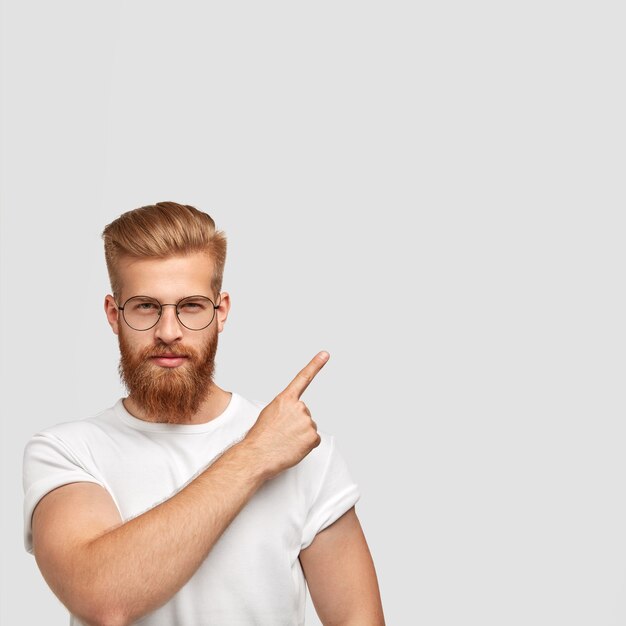 Un homme séduisant au gingembre a une barbe épaisse, vêtu d'une tenue décontractée, a une expression sérieuse, pointe l'index dans le coin supérieur droit