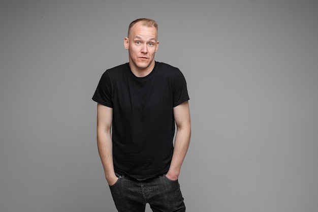 Homme séduisant aux cheveux courts et blonds portant un t-shirt noir et un jean gardant les mains dans les poches et surprenant de quelque chose