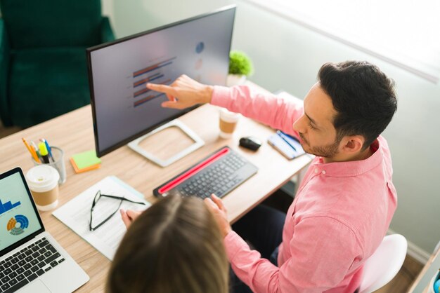 Homme séduisant pointant vers l'écran de l'ordinateur et la présentation de l'entreprise alors qu'il était assis au bureau. Collègues professionnels travaillant sur un rapport analytique