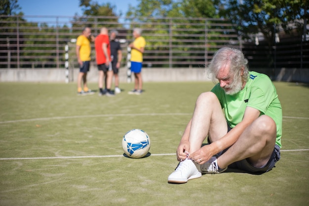 Homme senior assis sur des lacets de liage sur le terrain. Homme à la barbe grise en vêtements de sport se prépare pour l'entraînement. Football, sport, concept d'activités de loisirs